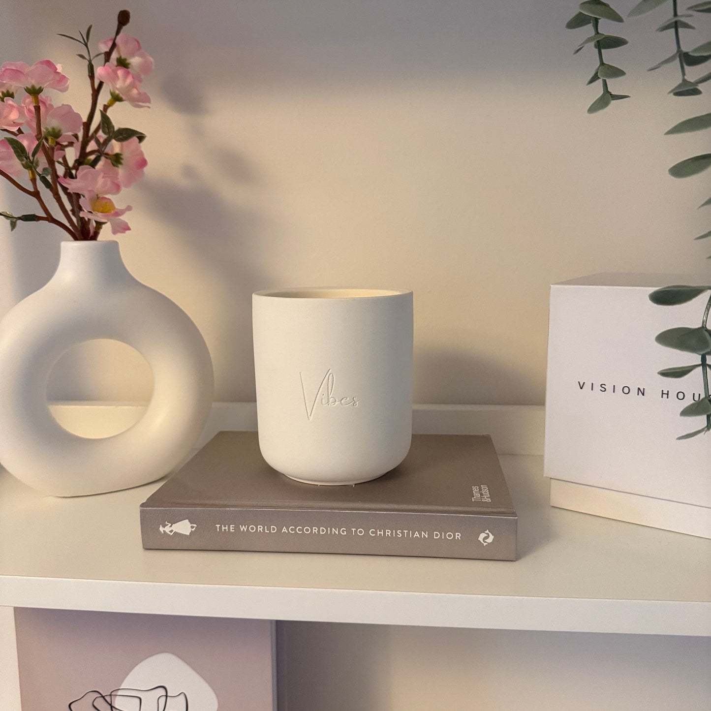 Decorative setup with a white ceramic candle, pink flowers, and a book on a shelf.