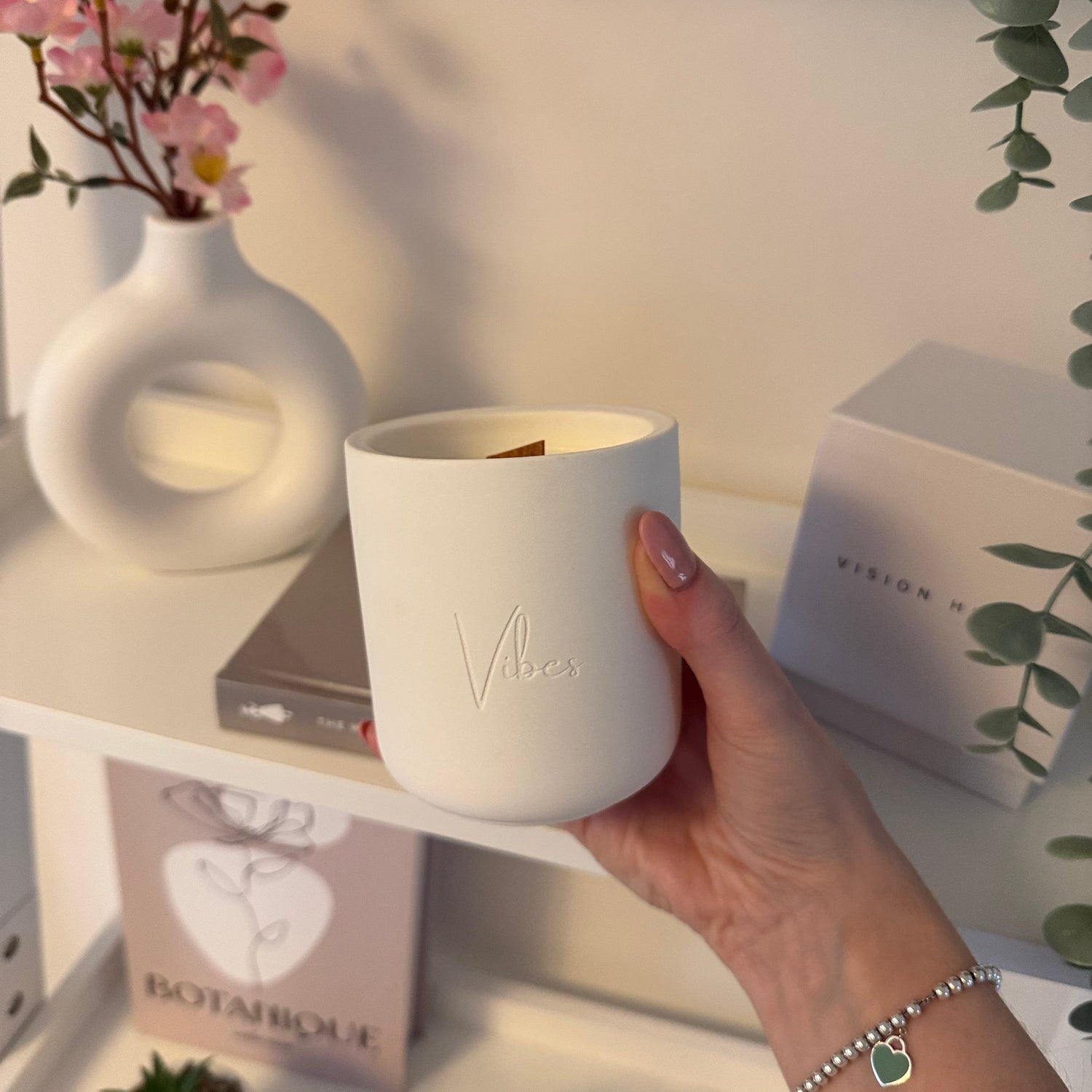 Hand holding a white ceramic candle with 'Vibes' branding, surrounded by books and plants on a light surface.