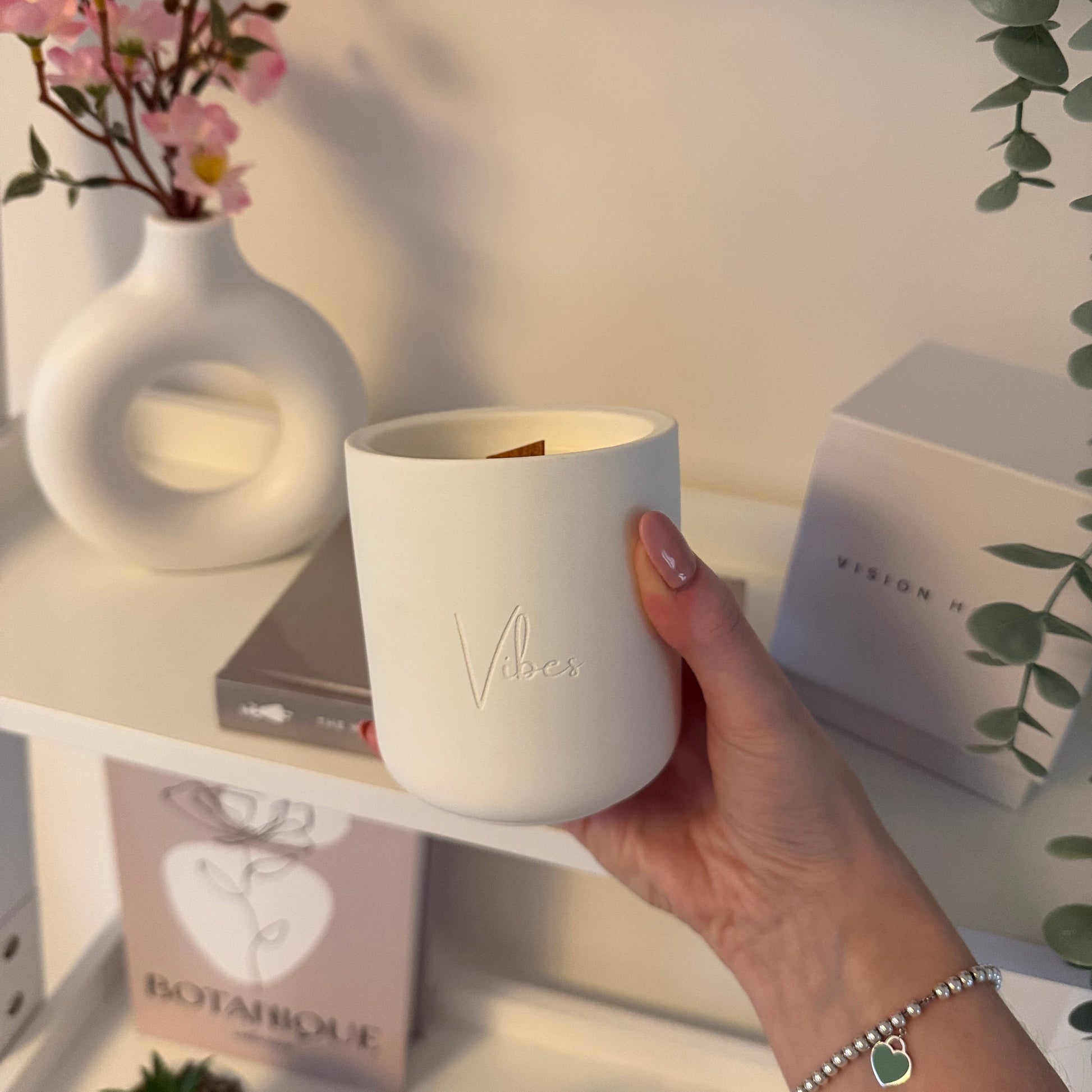 Hand holding a white ceramic candle with 'Vibes' branding, surrounded by books and plants on a light surface.